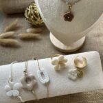 Sélection de bijoux artisanaux Nohulé Création sur présentoirs : collier avec pendentif fleur marron, bracelets fins avec breloques fleurs et géométriques, bague fleur blanche et bague d'oreille dorée.