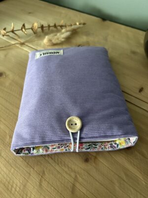 Pochette à livre artisanale matelassée Nohulé Création au motif velours mauve, fermée par un bouton en bois sur une table.