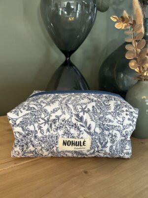 Fleuri bleu Trousse de toilette artisanale matelassée Nohulé Création au motif fleuri bleu et blanc, posée sur un meuble en bois devant un grand sablier.