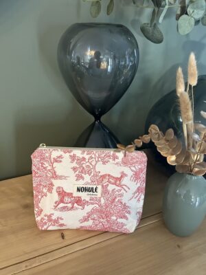 Trousse de maquillage artisanale Nohulé Création en toile de Jouy rouge et blanche, posée sur un meuble en bois devant un grand sablier décoratif.
