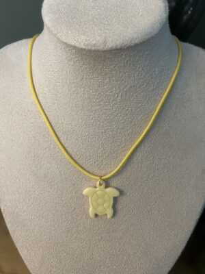 Collier ras de cou avec cordon de couleur jaune et pendentif artisanal en forme de petite tortue jaune clair Nohulé Création présenté sur un buste d'exposition blanc.